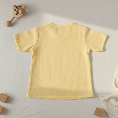 Yellow baby t-shirt on a light gray surface with wooden toys and a plant.